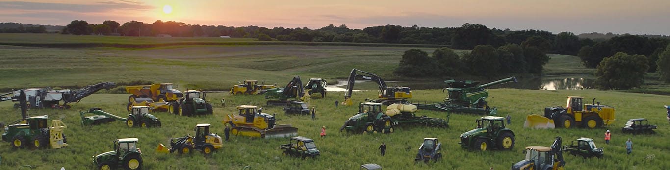 Geniş bir tarlada çeşitli John Deere ekipmanları arasında yürüyen insanlar.