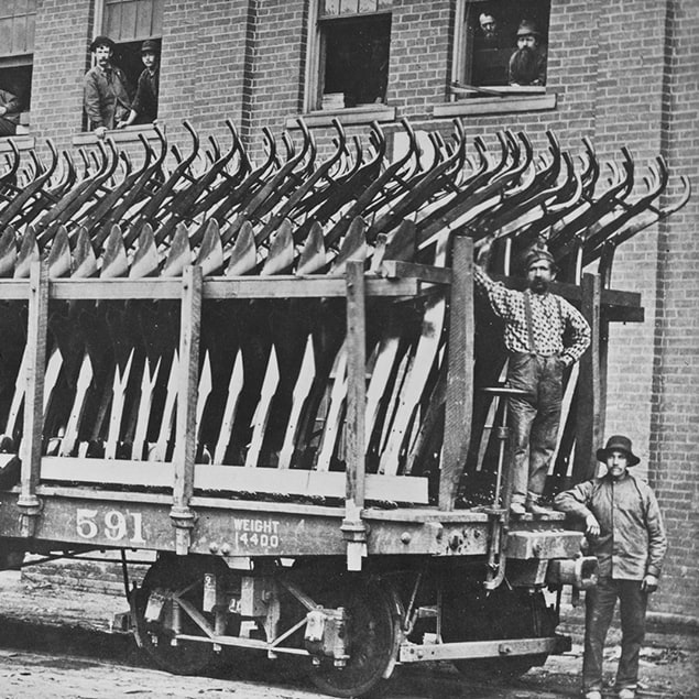 1882 yılından, nakledilmeye hazır çelik pulluklarla yüklü Deere & Co vagonunun yanında ayakta duran üç adamın ve arkadaki binanın penceresinden bakan fabrika çalışanlarının fotoğrafı