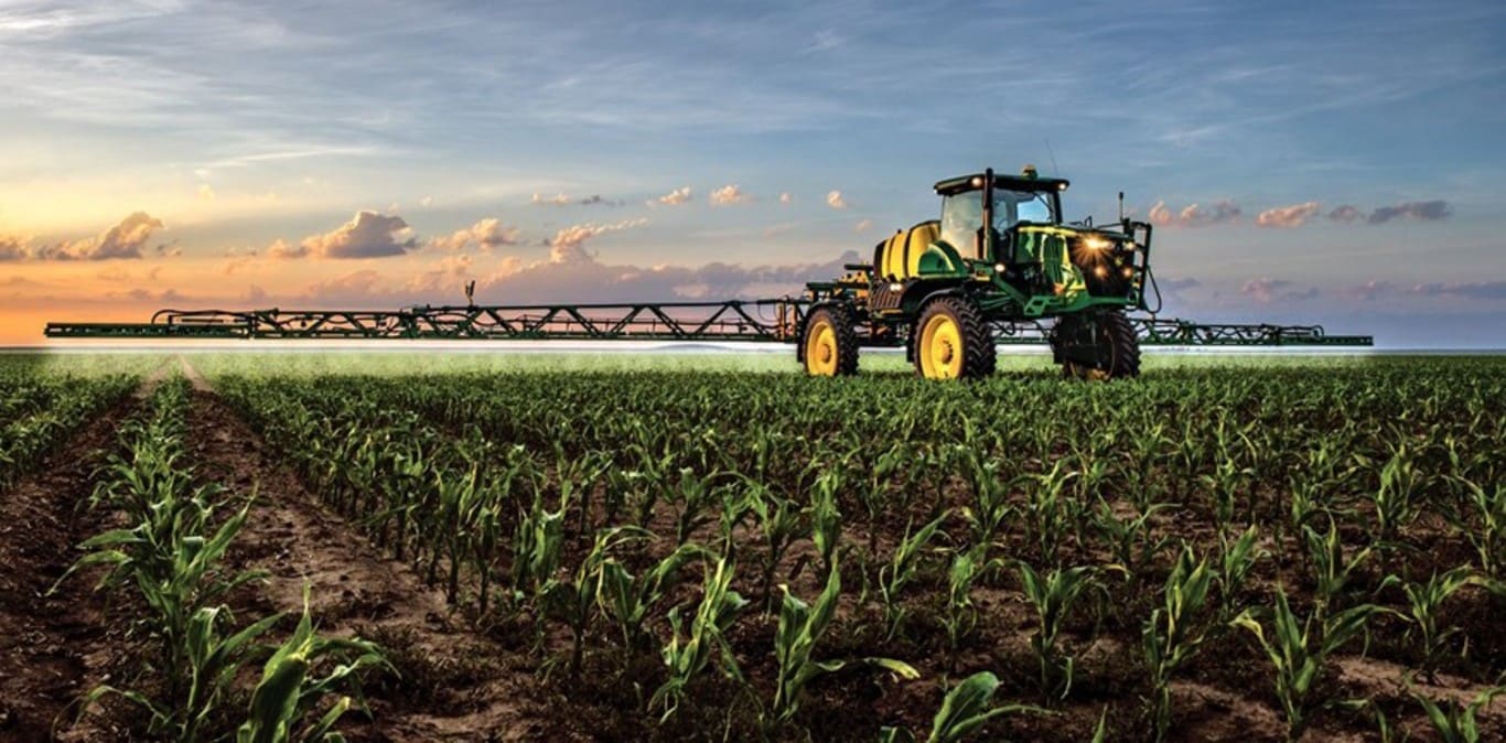 Arka planda gün batımı, bir tarlada John Deere ilaçlama makinesi çalışıyor