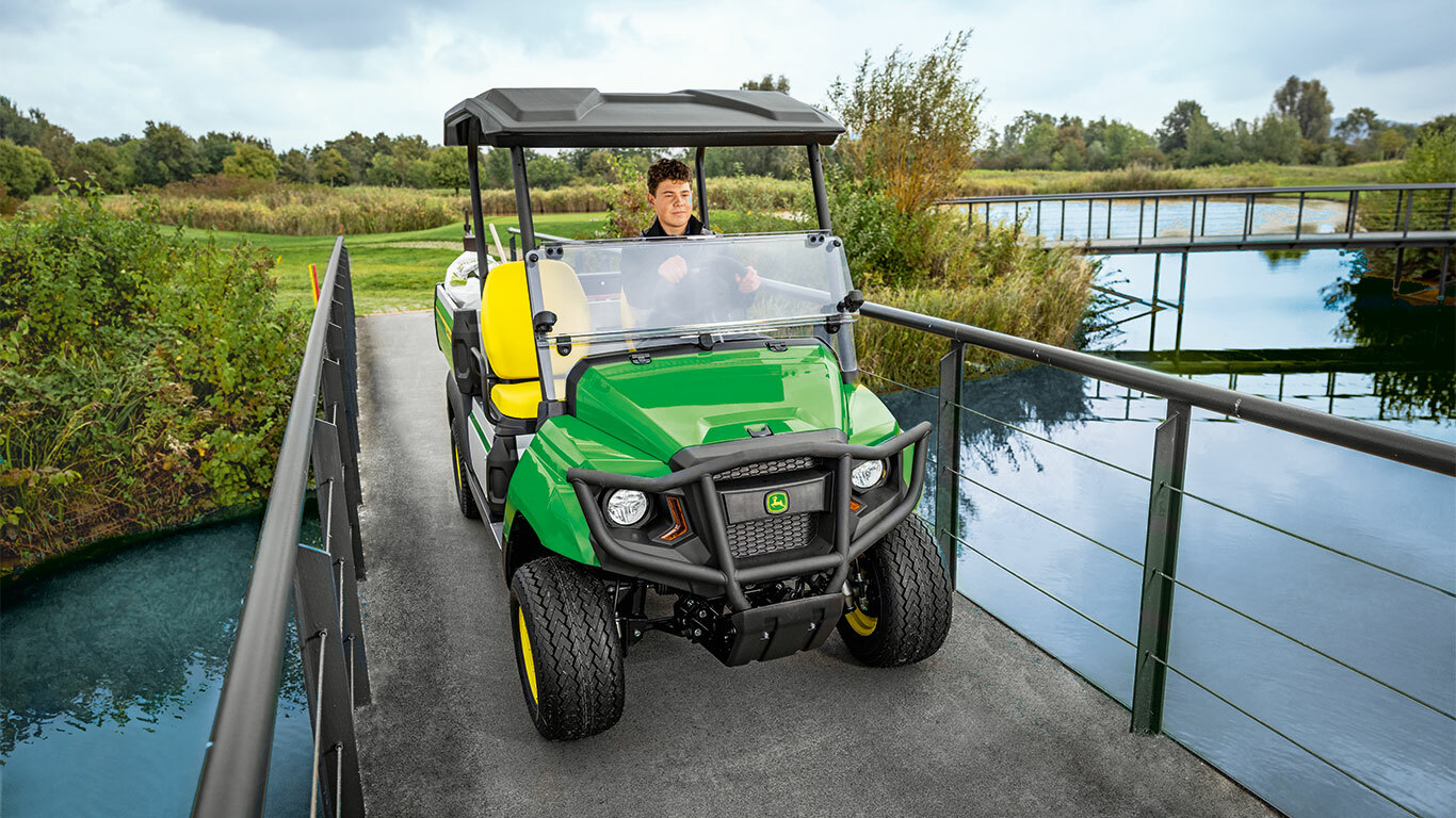 Yemyeşil ve ağaçlarla çevrili sakin bir su yolu üzerinde dar bir köprüden geçen John Deere Gator GS hizmet aracı. Yemyeşil ve ağaçlarla çevrili sakin bir su yolu üzerinde dar bir köprüden geçen John Deere Gator GS hizmet aracı.