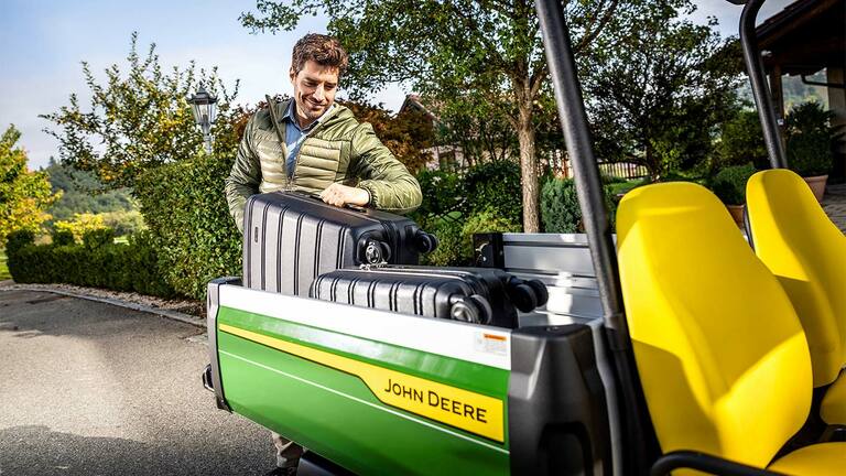 Yeşil ve ağaçlarla çevrili alanda, parlak sarı koltuklu John Deere Gator GS kargo bölümüne büyük siyah bir bavul yükleyen biri.
