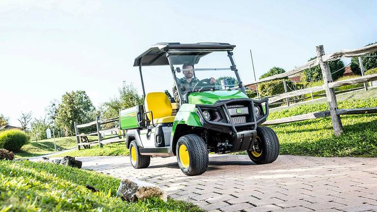 Çim ve ahşap çitlerle peyzajlı bir arazide asfaltlı bir yolda giden yeşil John Deere Gator GS hizmet aracı.