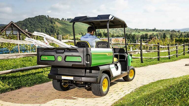 Arka planda manzaralı tepeler ve üzüm bağlarıyla tuğla yolda giden bir John Deere Gator GS'nin arkadan görünümü.