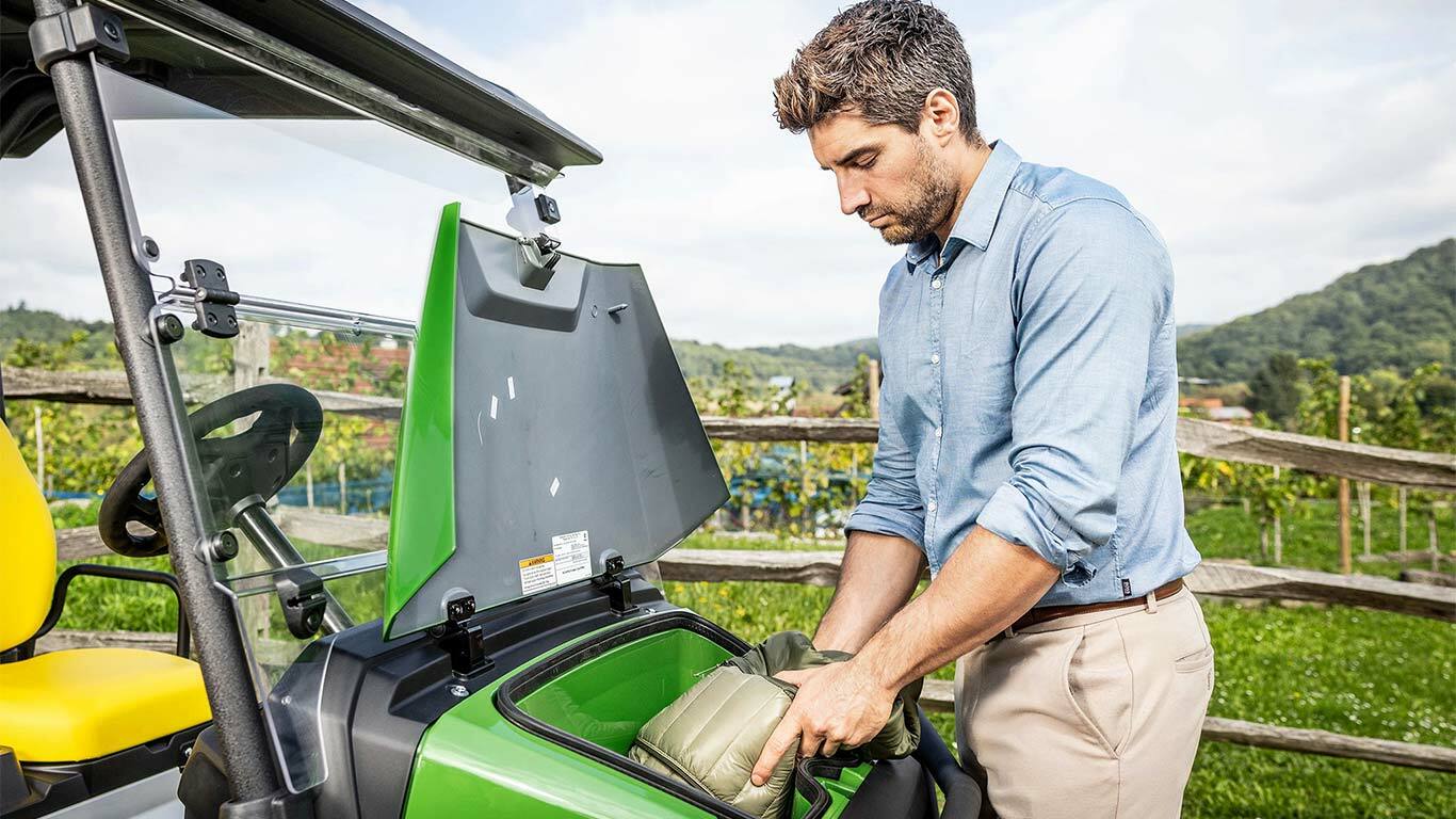 Ahşap bir çitin ve üzüm bağının yanında park edilmiş yeşil bir John Deere Gator GS'nin ön saklama bölmesine bir torba yerleştiren biri.