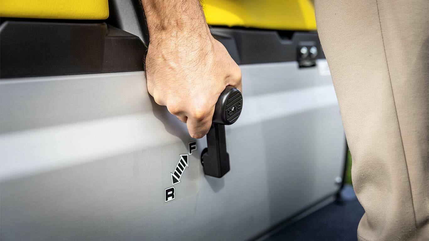 Sarı koltukların yanında ileri ve geri konumlarını gösteren vites değiştirme kolunu bir John Deere Gator GS üzerinde çalıştıran elin yakından görünümü.