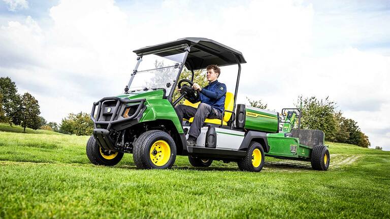 Kısmen bulutlu bir gökyüzünün altındaki çimen tarlasında ekipman çeken yeşil John Deere Gator elektrikli hizmet aracı.
