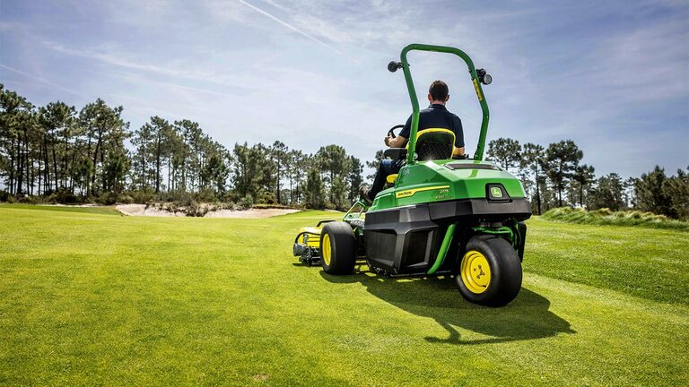 Bir kişi, yeşil bir John Deere 2775 E-Cut™ Elektrikli çim biçme makinesini, oldukça bakımlı bir golf sahasında, etrafta ağaçlar ve arka planda kısmen bulutlu bir gökyüzü varken sürüyor.