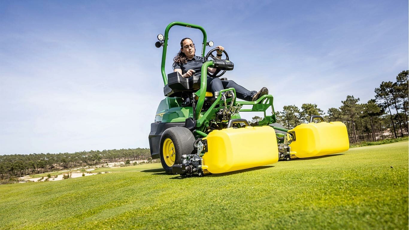 Bir kişi, yeşil ve sarı bir John Deere 2775 E-Cut™ Elektrikli çim biçme makinesini sürüyor, ön makaralar, golf sahası yeşili üzerinde sarı dış gövdeyle kaplı, ağaçlar var ve arka planda açık mavi gökyüzü var.