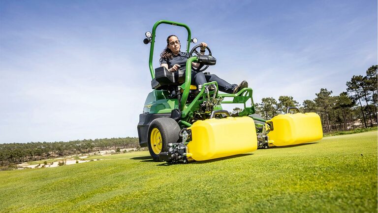 Bir kişi, yeşil ve sarı bir John Deere 2775 E-Cut™ Elektrikli çim biçme makinesini sürüyor, ön makaralar, golf sahası yeşili üzerinde sarı dış gövdeyle kaplı, ağaçlar var ve arka planda açık mavi gökyüzü var.