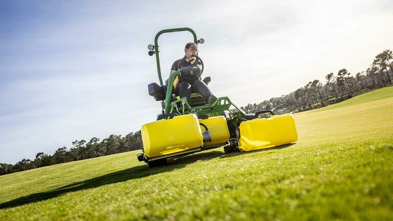 Bir kişi, yeşil ve sarı bir John Deere 2775 E-Cut™ Elektrikli çim biçme makinesini sürüyor, üç adet büyük ön makara, golf sahası yeşili üzerinde sarı dış gövdeyle kaplı, ağaçlar var ve arka planda parçalı bulutlu gökyüzü var.