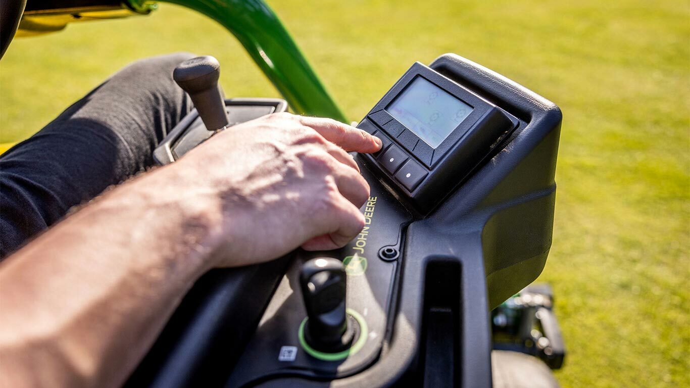 Bir John Deere 2775 E-Cut™ Elektrikli çim biçme makinesindeki TechControl panelini kullanarak, dijital ekranda düğmelere basan bir kişinin elinin yakından görünümü, joystick ve diğer kumandalar görülüyor.
