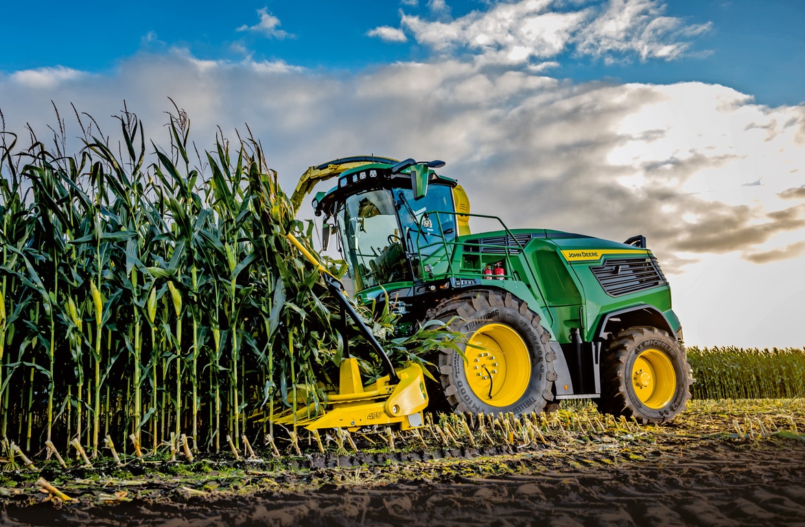 Mavi, bulutlu bir gökyüzünün altında tarlada mısır hasat eden John Deere silaj makinesi.