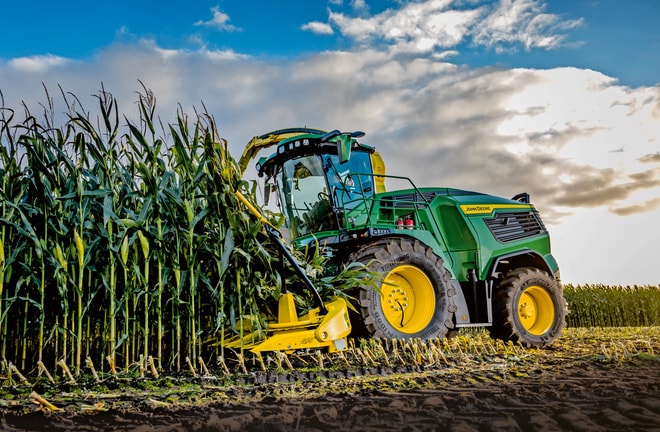 Mavi, bulutlu bir gökyüzünün altında tarlada mısır hasat eden John Deere silaj makinesi.