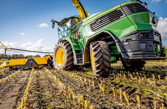 Hasat edilmiş bir mısır tarlasında çalışan John Deere silaj makinesi, makinenin büyük lastiklerini ve kesme mekanizmasını berrak mavi bir gökyüzüne karşı sergiliyor.