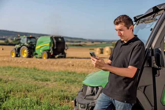 Arka planda bir John Deere VR balya makinesi ile cep telefonundaki balya belgeleme verilerini kontrol eden kişi